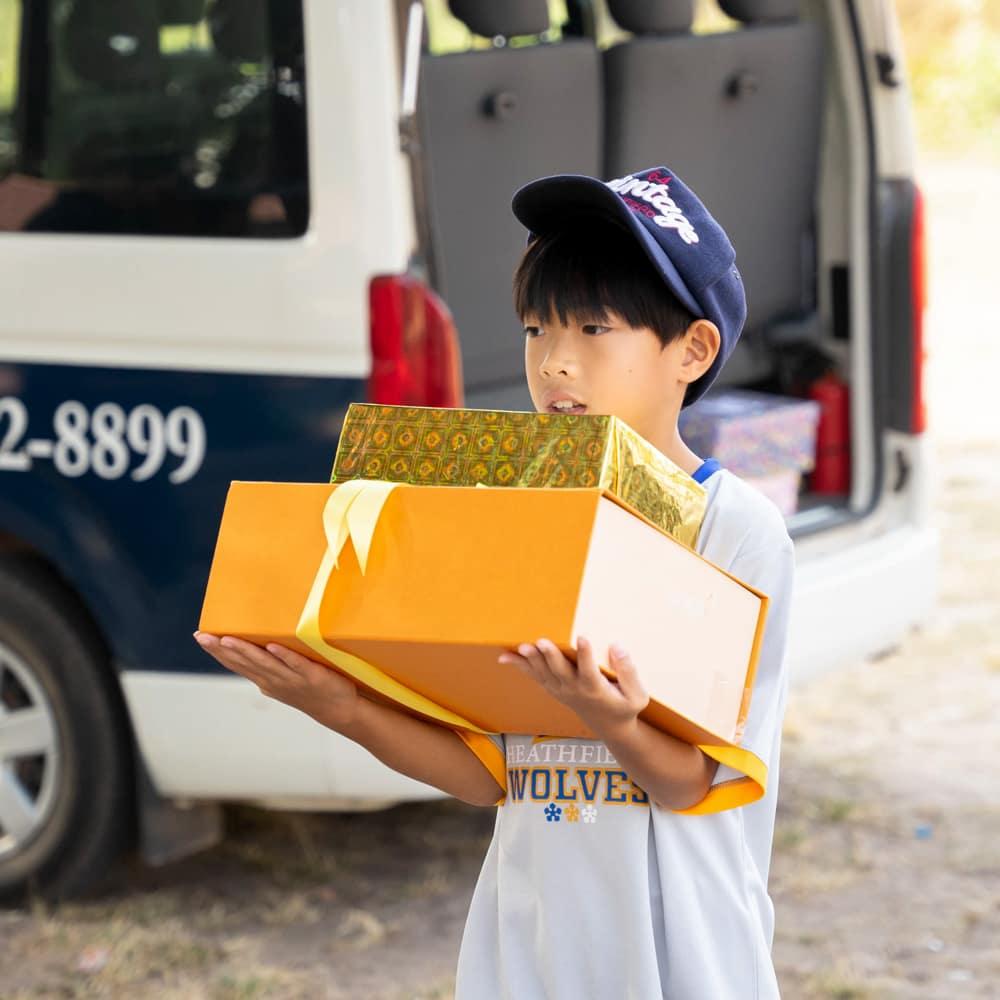 A student holding two brightly wrapped gift boxes near the school van during the Holiday Box Project.