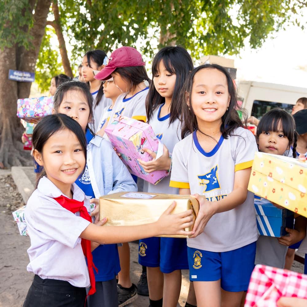 Children from the school handing over wrapped gift boxes to students from another school under the shade of trees.