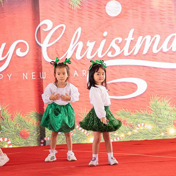 Two students in festive green skirts and white tops perform a cheerful Christmas-themed dance on stage.