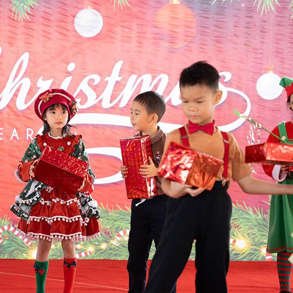 Children in festive costumes hold shiny red gift boxes as they perform a cheerful Christmas-themed dance.