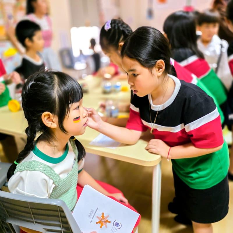 Two young girls painting each other's faces in a classroom during International Day celebrations.