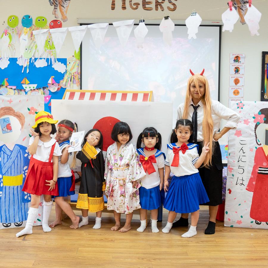 A group of students dressed in Sailor Moon-themed costumes and a yukata, posing with their teacher in front of a Japan-themed classroom display.