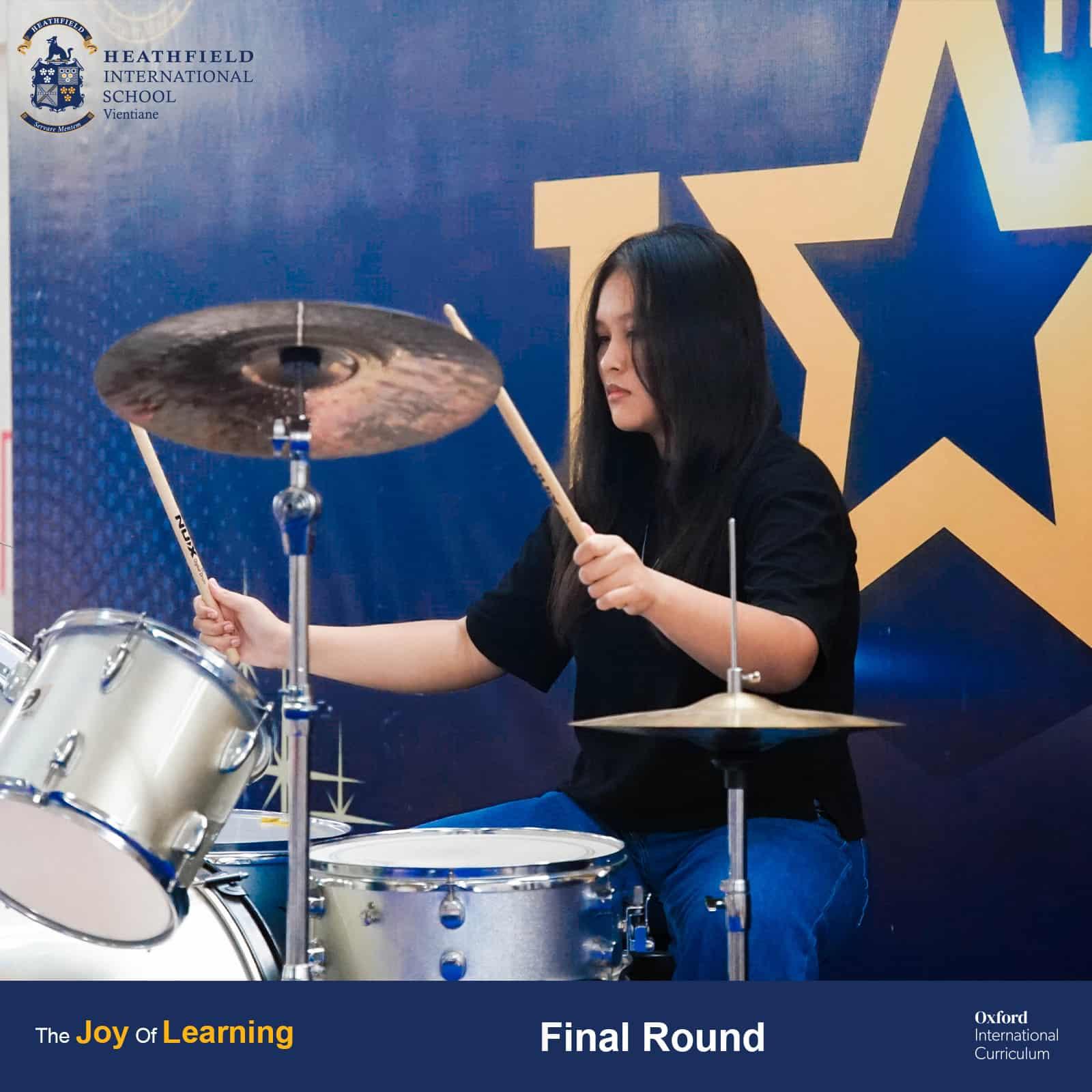 A drummer energetically performing on stage as part of a three-person rock band during Heathfield's Got Talent finals.